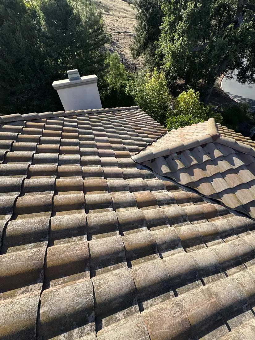 Finished barrel tile roof with a hip run and chimney, hillside behind it.